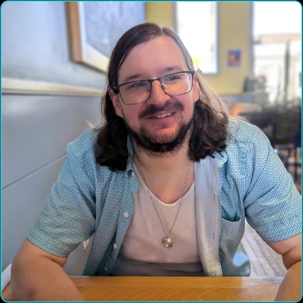 A man sitting at a table and facing the camera while his eyes are looking to the right. He has a slight grin. He's wearing a white tank top with a light blue unbuttoned shirt over it. He has a necklace with a silver ring hanging from it. His background is a cafe.